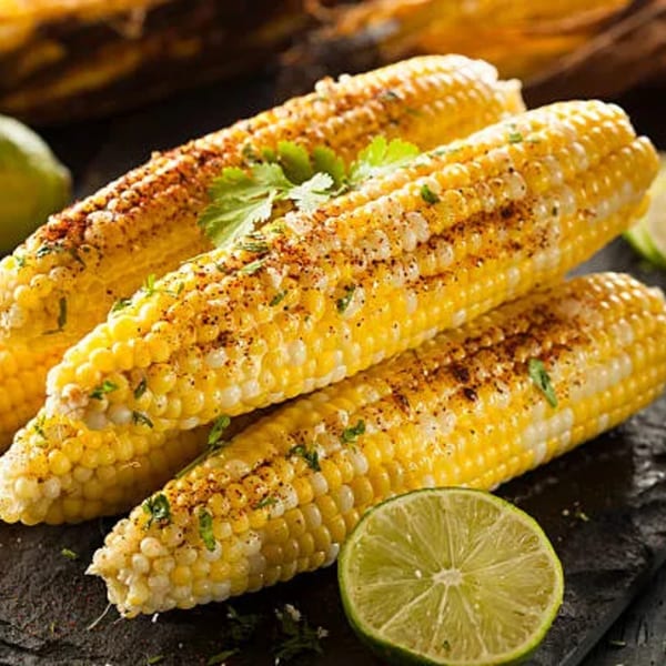 Three ears of grilled corn on the cob, seasoned with spices and cilantro, with lime wedges.