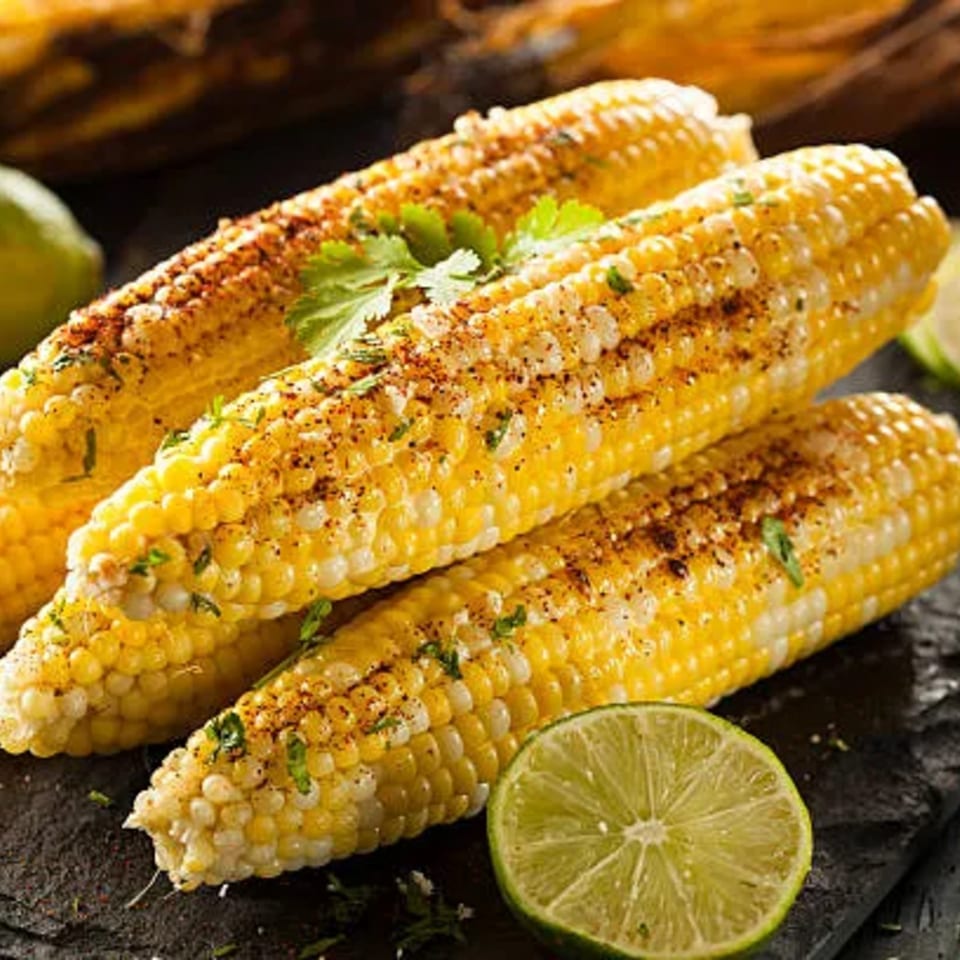 Three ears of grilled corn on the cob, seasoned with spices and cilantro, with lime wedges.