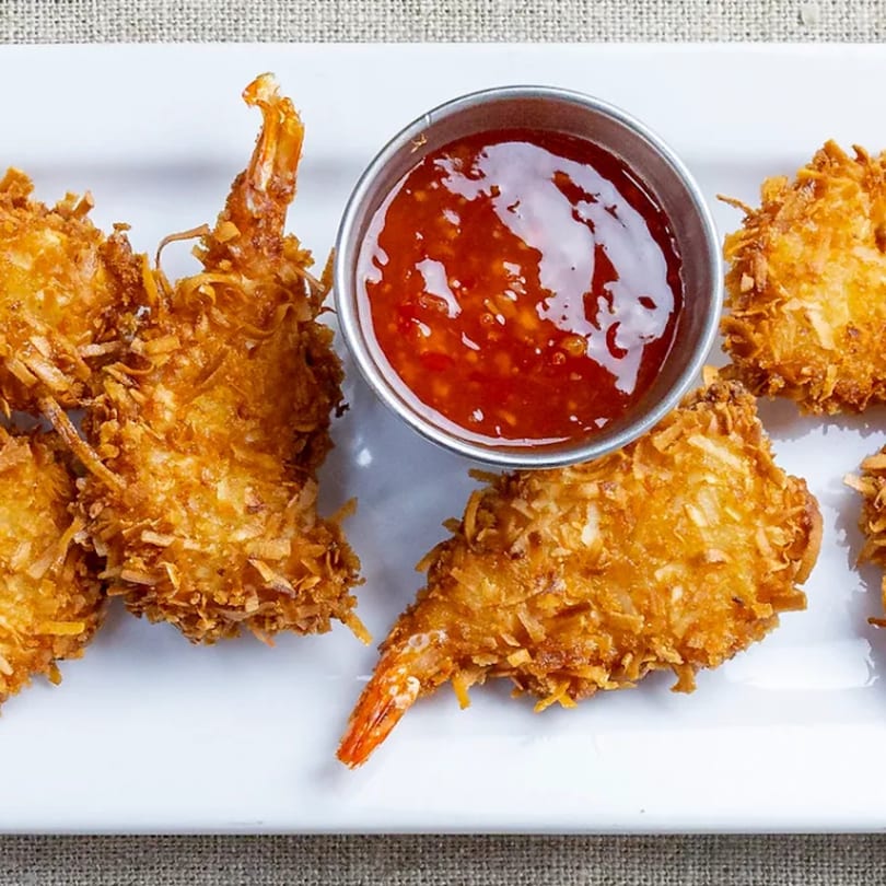 Crispy golden-brown coconut shrimp with a side of sweet chili dipping sauce on a white plate.