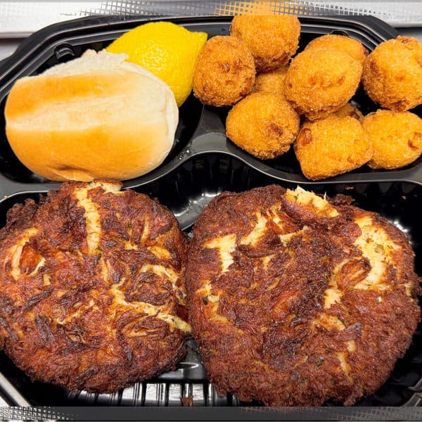 Two crab cakes, hush puppies, a lemon wedge, and a dinner roll in a black to-go container.