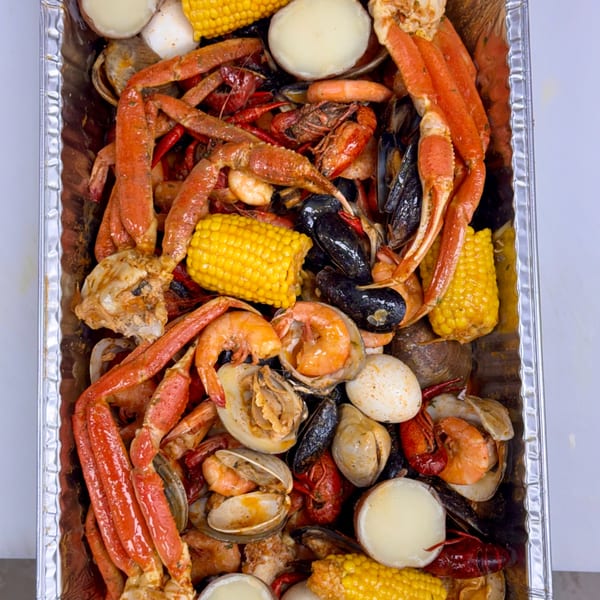 A large foil tray filled with a seafood boil including crab legs, shrimp, mussels, corn, and potatoes.