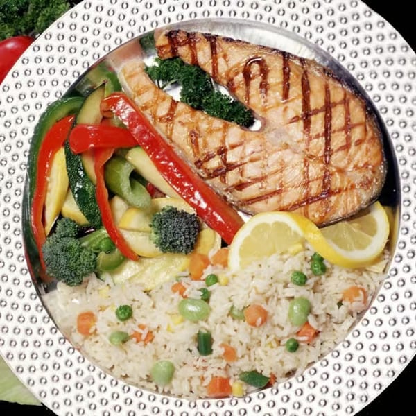 Grilled salmon steak with mixed vegetables, rice pilaf, and lemon slices on a decorative plate.