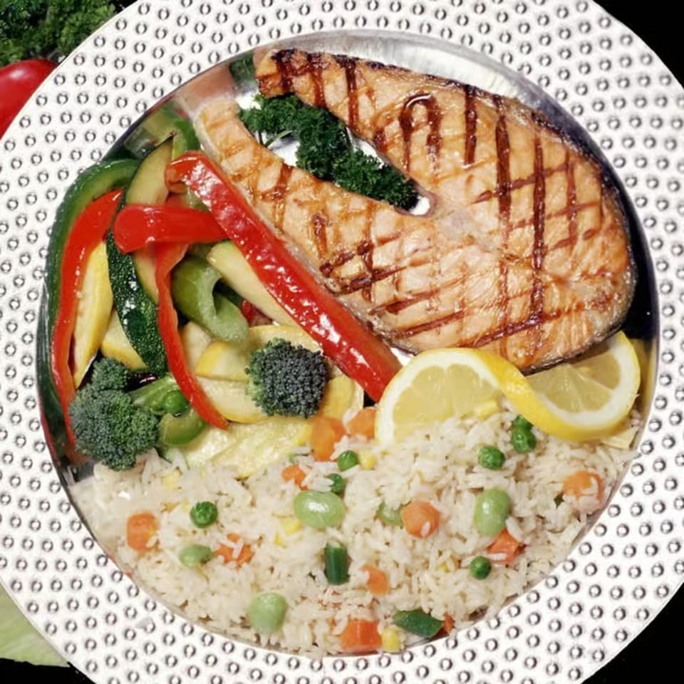 Grilled salmon steak with mixed vegetables, rice pilaf, and lemon slices on a decorative plate.