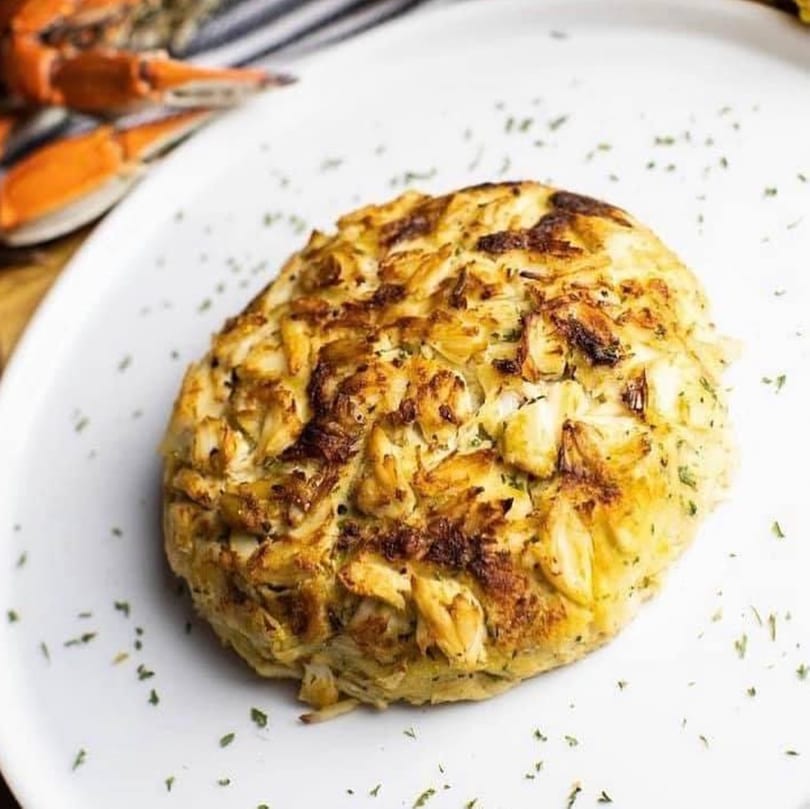 A golden-brown Cameron's Seafood crab cake, seasoned with herbs, sits on a white plate.
