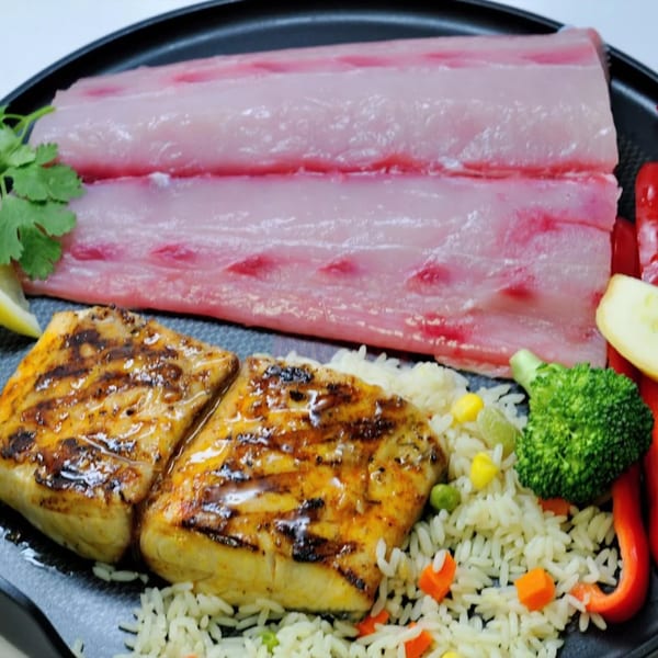 Two grilled fish fillets with rice and vegetables next to raw fish fillets on a black plate.