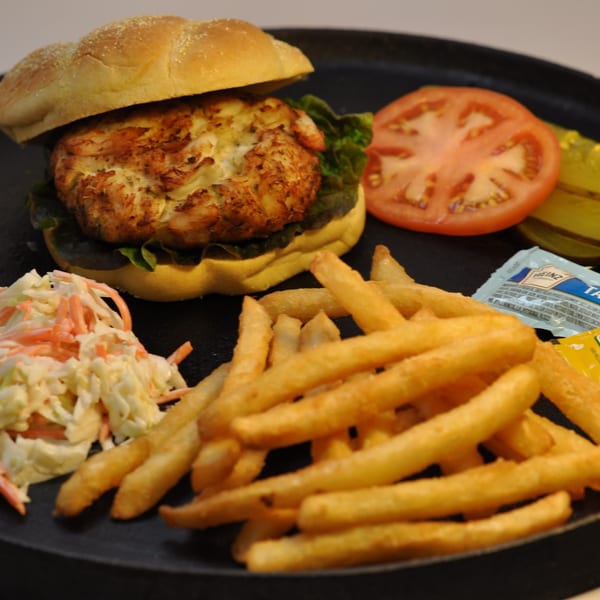 Crab cake sandwich with lettuce, tomato, pickles, coleslaw, and french fries on a black plate.