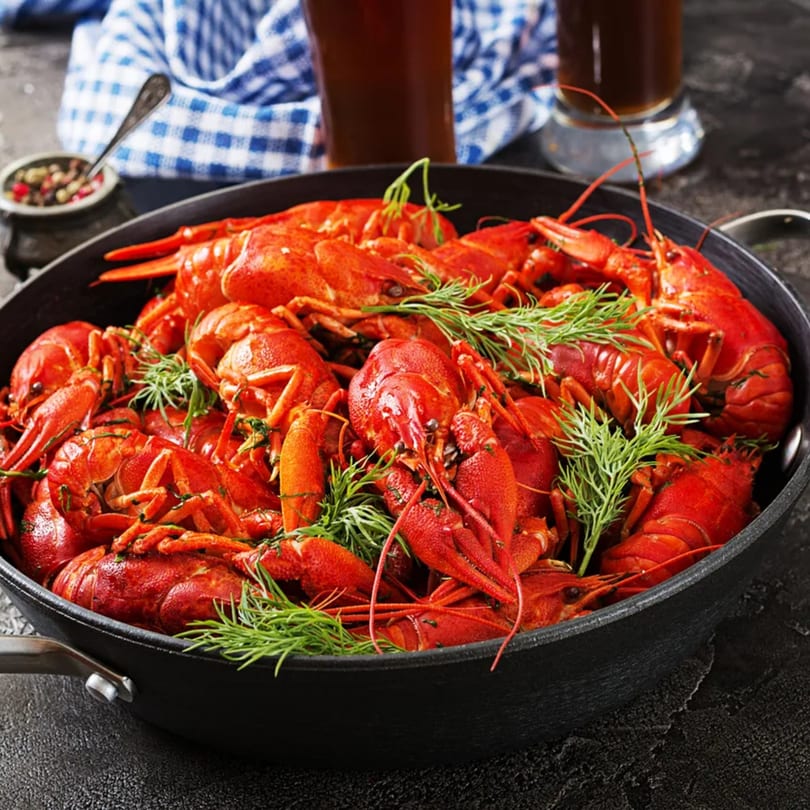 A black pan filled with bright red cooked crawfish and fresh dill, with two glasses of beer.
