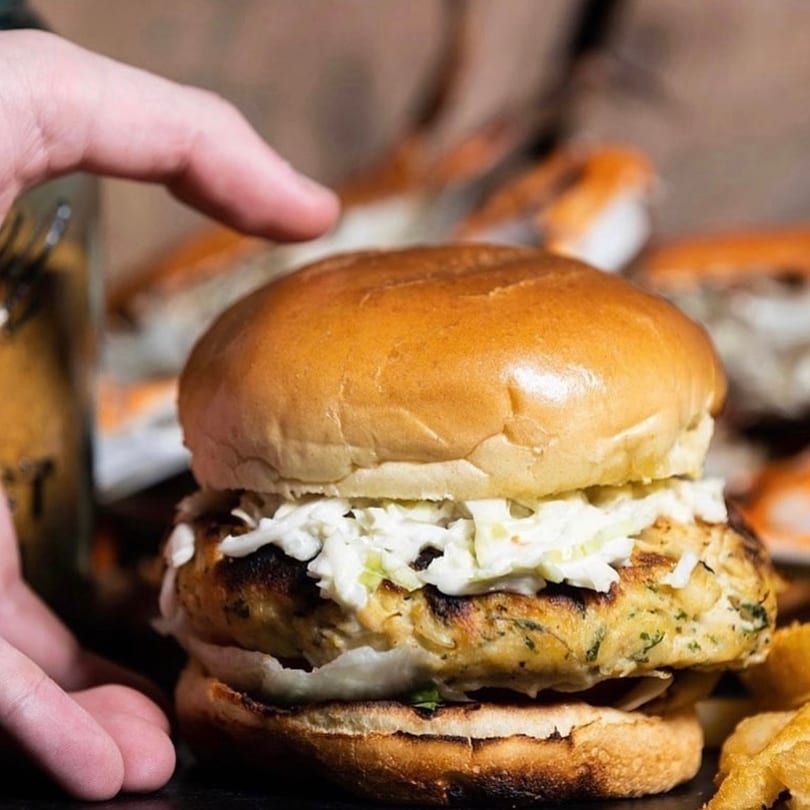 A hand reaches for a Cameron's Seafood crab cake sandwich topped with coleslaw and fries.