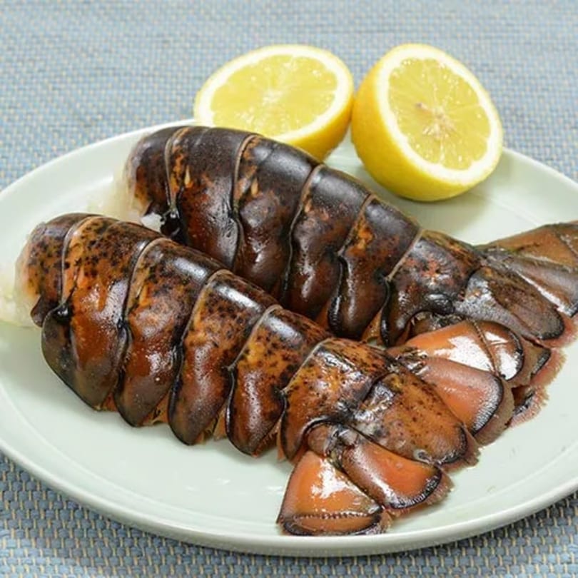 Two raw lobster tails with two lemon halves on a white plate, ready for cooking.